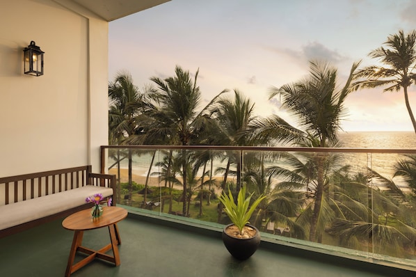 A balcony with a view of the beach and palm trees, a wooden bench, a small table with a vase, and a potted plant.