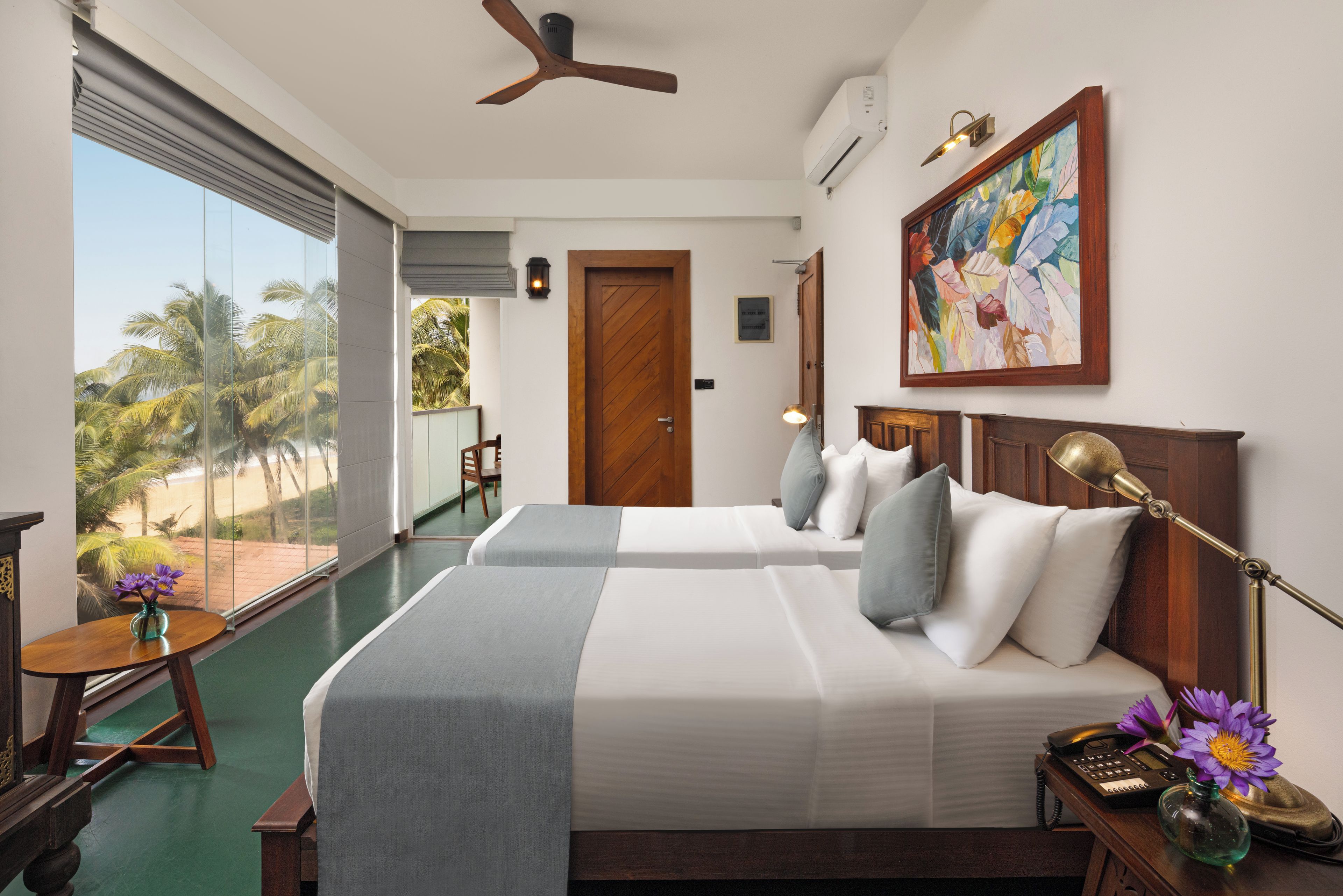 A hotel room with two beds, a desk, a chair, a view of palm trees, and a painting on the wall.