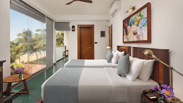 A hotel room with two beds, a desk, a chair, a view of palm trees, and a painting on the wall.