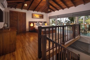 Una habitación con piso de madera, una cama con almohadas, una mesita de noche con una lámpara, una puerta de madera y una ventana con vista a palmeras.