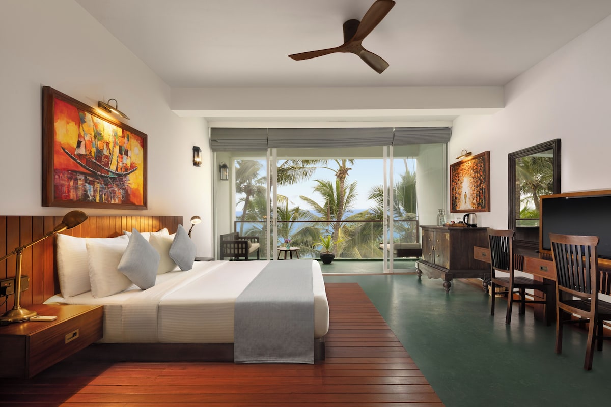 A hotel room with a bed, a desk, a chair, a ceiling fan, and a view of palm trees and the ocean.