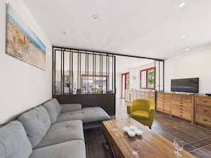 A modern living room with a sectional sofa, a wooden coffee table with wine glasses, a yellow armchair, and a TV on a wooden cabinet. - Family house for 8 – garden & BBQ (Longeville-sur-Mer)