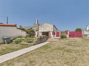 A stone building with a church steeple in the background, a paved pathway, and a fenced area with a small garden. - Family house for 8 – garden & BBQ (Longeville-sur-Mer)