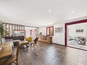 A spacious living area with a wooden floor, a sofa, a dining table, and a kitchen area visible through an open door. - Family house for 8 – garden & BBQ (Longeville-sur-Mer)