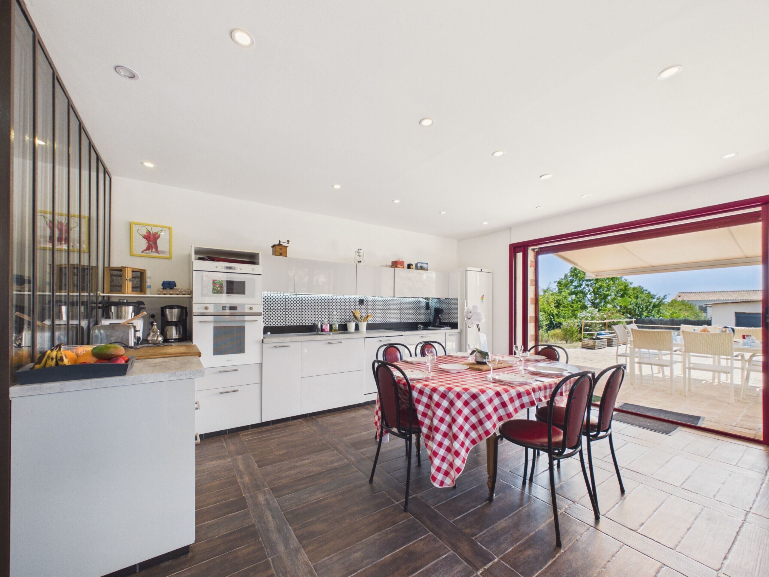 A modern kitchen with white cabinets, a dining area with a checkered tablecloth, and a view through a glass door to an outdoor patio.