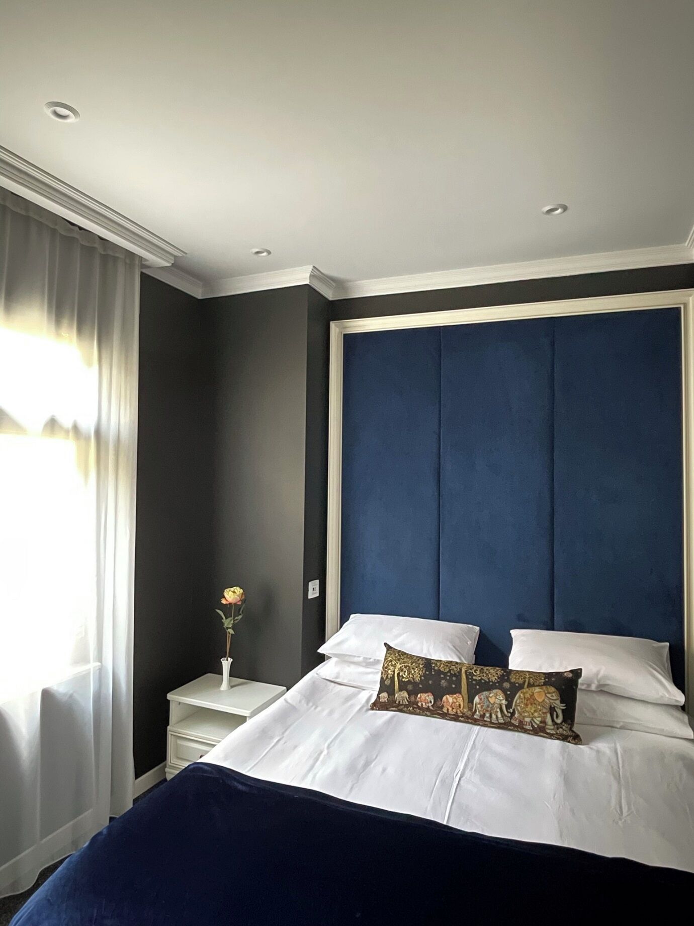 Ein Schlafzimmer mit einem großen Bett, weißer Bettwäsche, einem dunkelblauen Kopfteil, einem Nachttisch mit Blumenvase und einem Fenster mit durchsichtigen Vorhängen.
