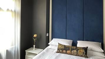 Ein Schlafzimmer mit einem großen Bett, weißer Bettwäsche, einem dunkelblauen Kopfteil, einem Nachttisch mit Blumenvase und einem Fenster mit durchsichtigen Vorhängen.