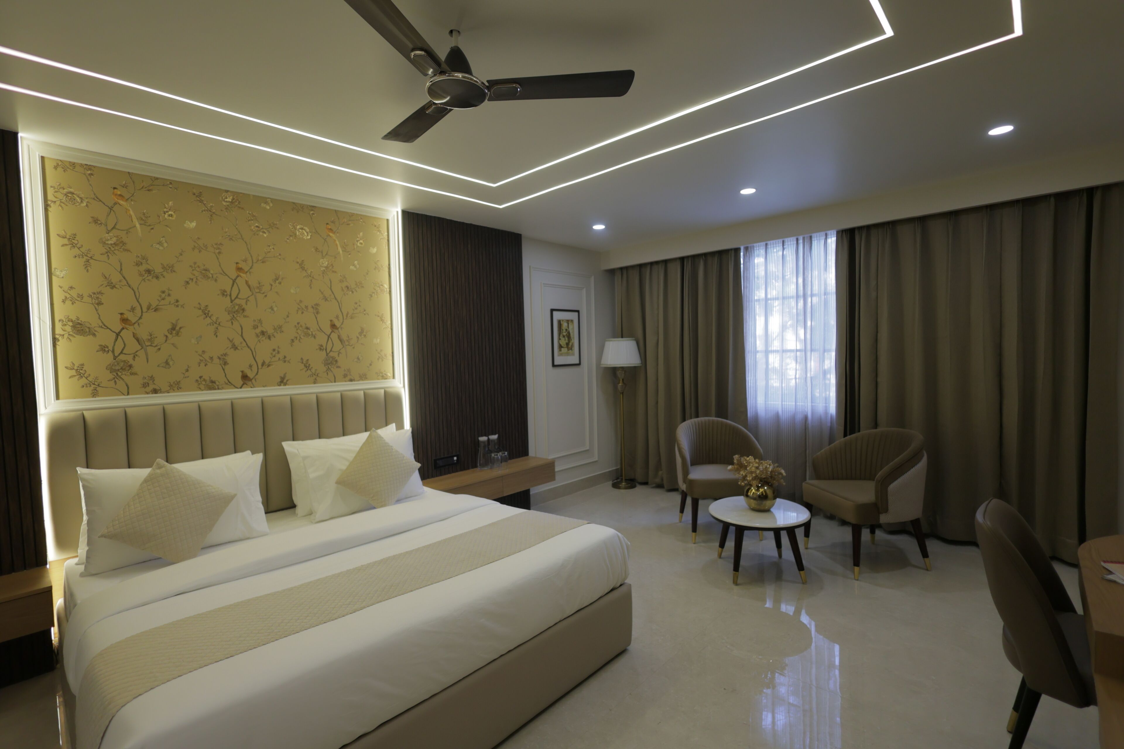 A hotel room with a large bed, two armchairs, a small table, a ceiling fan, and a wall with floral wallpaper.