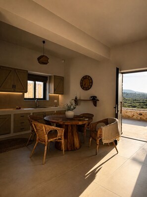 A spacious kitchen with wooden cabinets, a round dining table, wicker chairs, and a view through an open door. - Conte Maris Tower III - Sea View Mediterranean Escape (Tragaki)