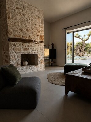 A modern living room with a stone fireplace, a sofa, a coffee table, and a view of the outdoors. - Conte Maris Tower III - Sea View Mediterranean Escape (Tragaki)