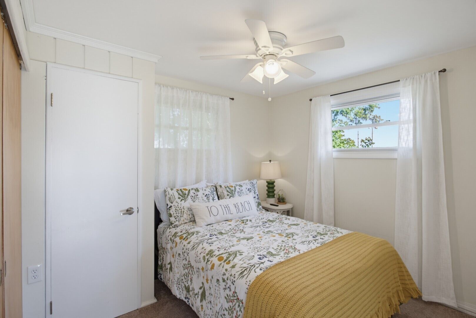 Un dormitorio con cama, ventilador de techo, mesita de noche con lámpara, ventana con cortinas y una puerta.