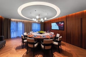 Ein Konferenzraum mit rundem Tisch, Stühlen, einer Chandelierleuchtre, einem Fernsehbildschirm und einem großen Fenster mit Vorhängen.