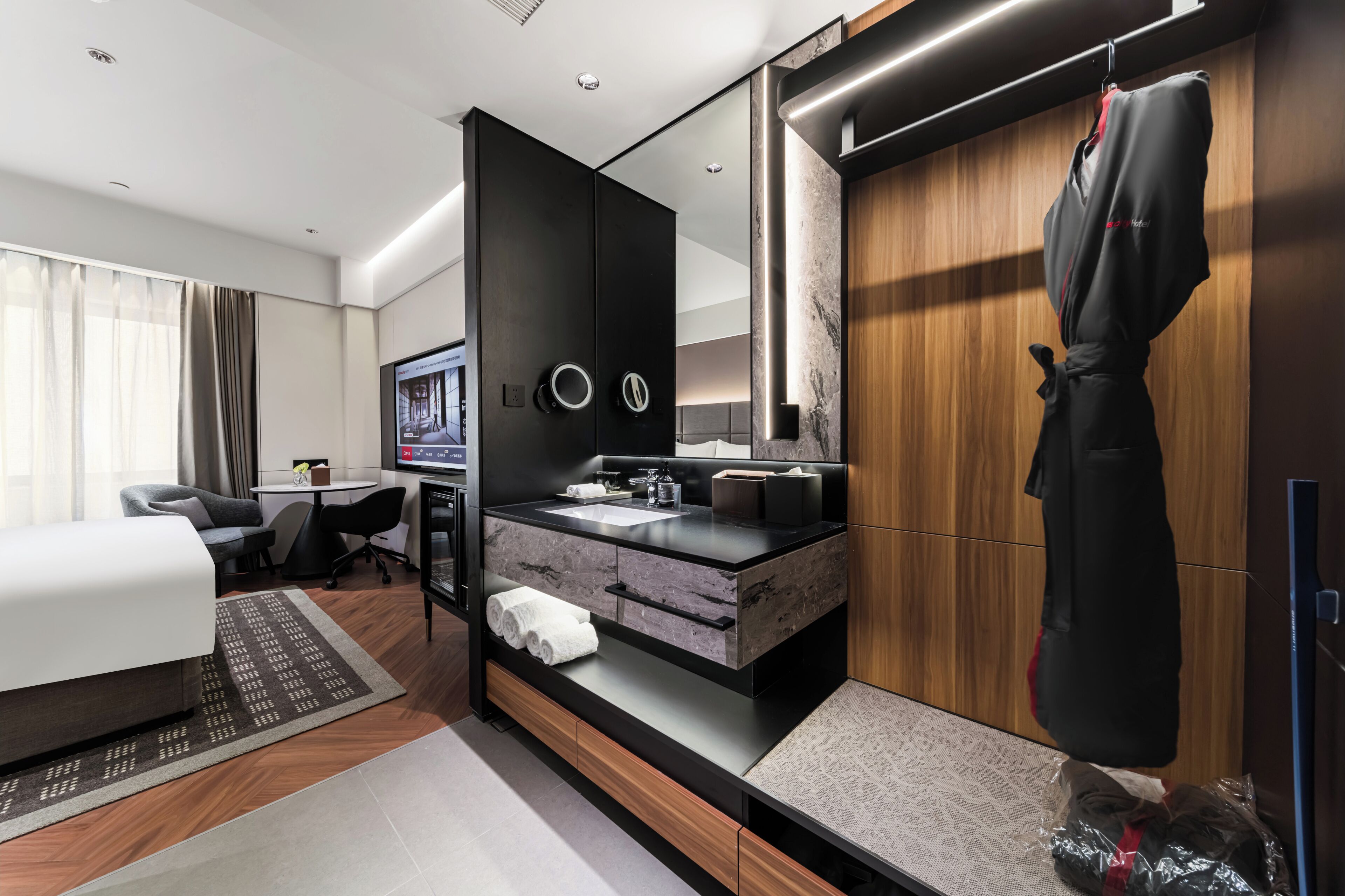 A modern hotel room with a bed, a desk, a bathroom with a vanity, and a robe hanging by the door.