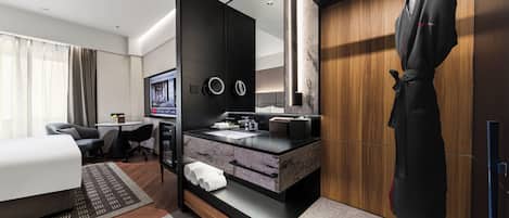 A modern hotel room with a bed, a desk, a bathroom with a vanity, and a robe hanging by the door.