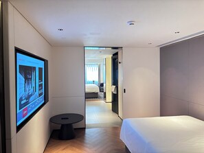 Una habitación de hotel moderna con una cama, una mesita redonda pequeña, un televisor montado en la pared y vistas a la habitación contigua.