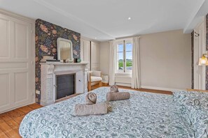 A bedroom with a bed, a fireplace, a window with curtains, and a view of greenery outside.