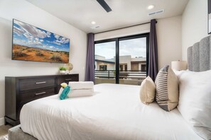A modern hotel room with a large bed, a flat-screen TV mounted on the wall, a dark wooden dresser, and a large window offering a view of the building's exterior. - Hurricane 3BR - Hot Tub & Mountain Views (Hurricane)