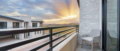 Ein Balkon mit Blick auf einen Sonnenuntergang über einer Berglandschaft, ein Korbstuhl und eine Steinmauer.