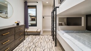 Un dormitorio moderno con una cama individual superpuesta, un aparador con cajones, un banco, un espejo grande y una ventana con vistas al exterior.