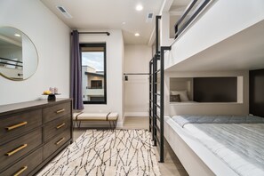 Ein modernes Schlafzimmer mit Etagenbett, einer Kommode mit Schubladen, einer Bank, einem großen Spiegel und einem Fenster mit Blick nach außen.