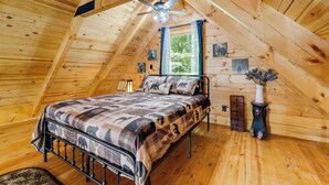 Un dormitorio acogedor en el ático, con cama, paredes de madera, ventilador de techo y ventana con cortinas.