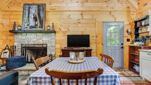 Un interior de cabaña acogedor con chimenea de piedra, televisor, mesa de comedor con sillas y estantería.