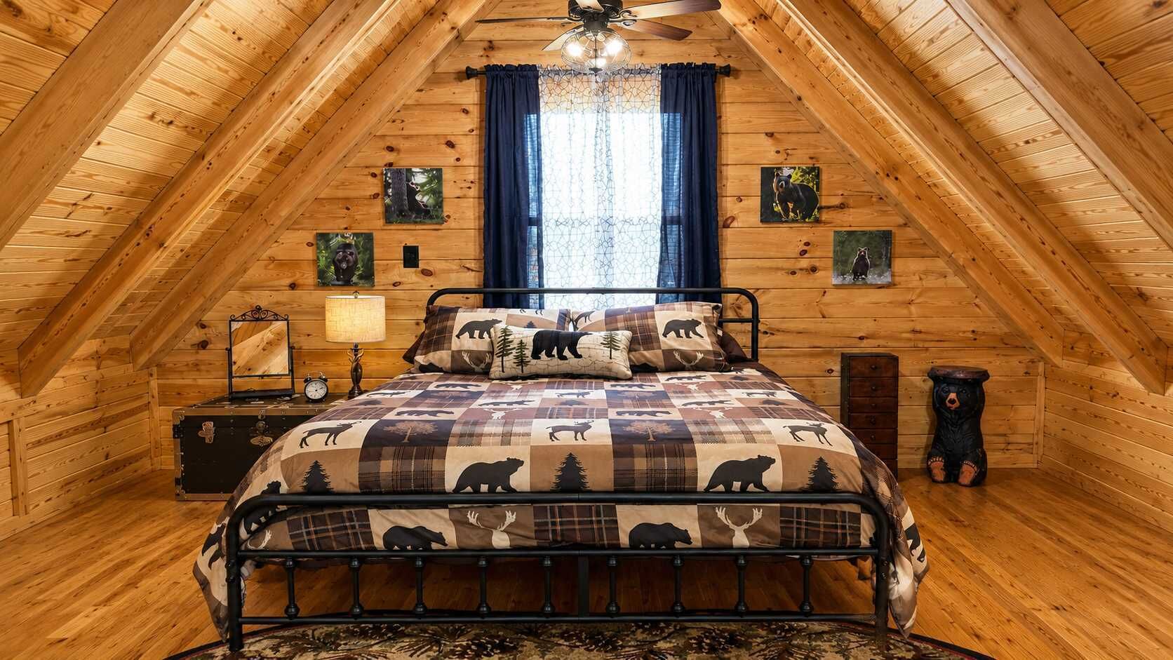 A cozy attic bedroom with a wooden sloped ceiling, a bed with a bear-themed quilt, a nightstand with a lamp, and a small bear statue.