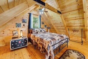 A cozy attic bedroom with a sloped ceiling, a bed with a patterned bedspread, a small bedside table with a clock, and a window with curtains.