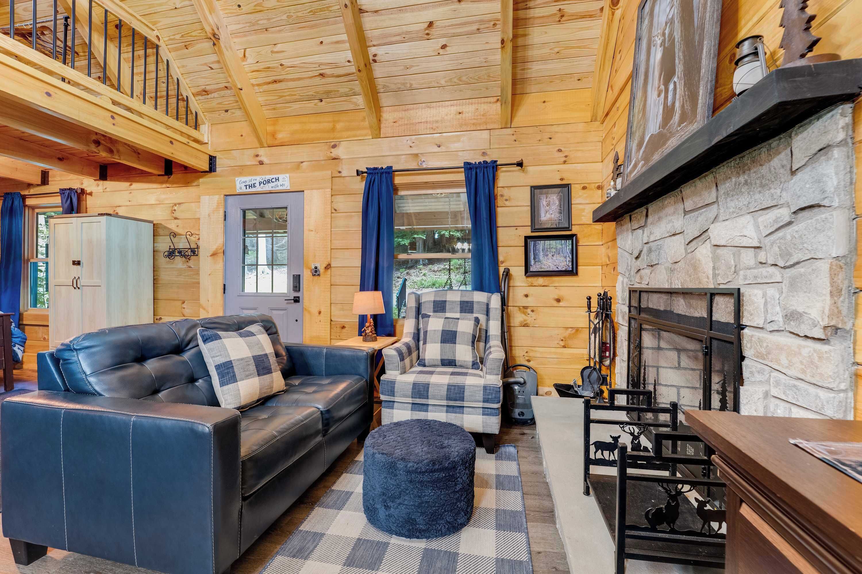 A cozy cabin interior with a leather sofa, plaid armchair, ottoman, stone fireplace, and a staircase leading to an upper level.