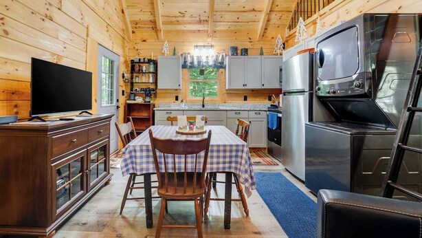 Interior de una cabaña de madera con una mesa de comedor, sillas, televisor, armarios de cocina, refrigerador y ventana con vistas a la vegetación.