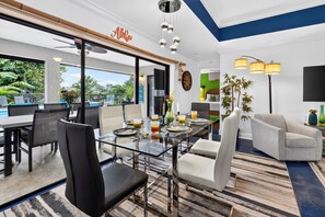 Habitación de hotel moderna con una mesa de comedor de vidrio, asientos cómodos, un ventanal con vistas y un televisor de pantalla plana.