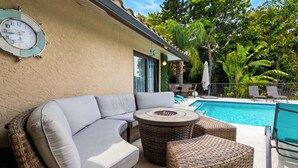 A poolside area with a wicker sofa, a round table, and a clock on the wall. - Private Pool + Spa + Outdoor Dining | Gulf Beaches | Blue Sunset (Bonita Springs)