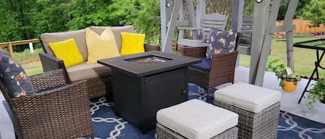 Un patio exterior con mobiliario de mimbre, una mesa negra y un tapete azul estampado.