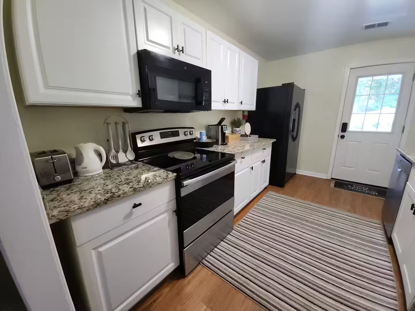 A kitchen with white cabinets, a black microwave, a stainless steel refrigerator, a granite countertop, and a striped rug.