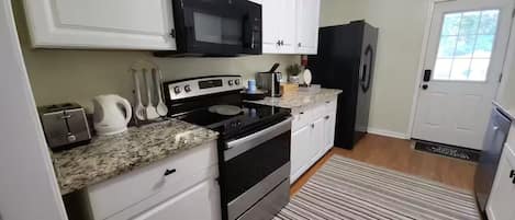 A kitchen with white cabinets, a black microwave, a stainless steel refrigerator, a granite countertop, and a striped rug.