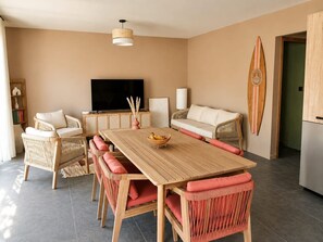 A modern living room with a wooden dining table, wicker furniture, a flat-screen TV, and a surfboard decoration. - Villa Mezzanotte in Le Brusc - 80 meters from the sea - Pool - Parking - Sleeps 8 - Cozy (Six-Fours-les-Plages)