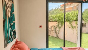Quarto com cama, almofadas, luminária, um quadro e vista para um jardim através de uma porta deslizante.