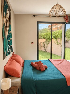 A bedroom with a bed, pillows, a lamp, a painting, and a view of a garden through a sliding door. - Villa Mezzanotte in Le Brusc - 80 meters from the sea - Pool - Parking - Sleeps 8 - Cozy (Six-Fours-les-Plages)