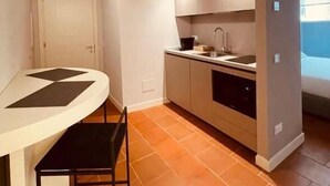 Una cocina compacta con encimera blanca, un lavamanos y un horno empotrado. Hay una puerta que da a otra habitación y una ventana que proporciona luz natural.