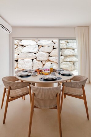 Un coin repas avec une table ronde dressée pour quatre personnes, entourée de chaises en osier, avec un mur de pierre et une grande fenêtre en arrière-plan.