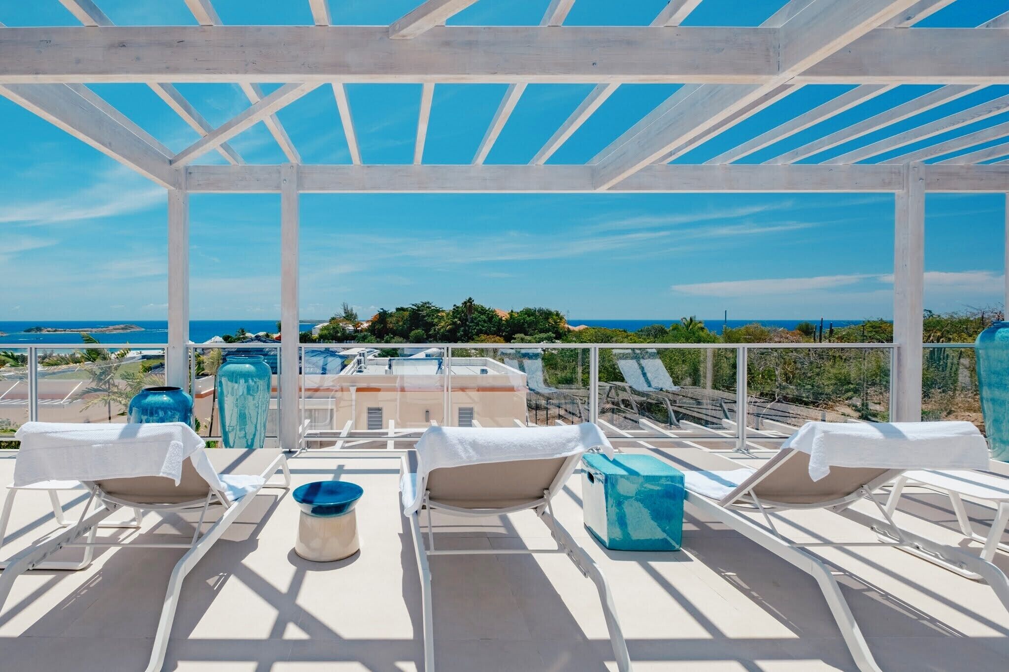 A rooftop terrace with white sun loungers, a blue vase, and a view of the ocean.