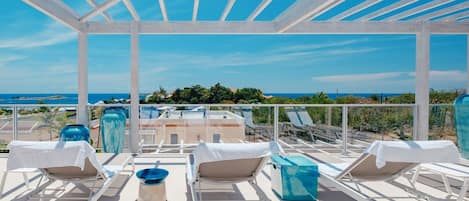 A rooftop terrace with white sun loungers, a blue vase, and a view of the ocean.