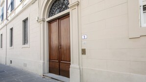 Fachada de edifício com uma grande porta dupla de madeira, uma pequena janela com persianas e uma placa com número na parede.