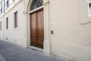Eine Gebäudefassade mit einer großen Holztür, einem kleinen Fenster mit Fensterläden und einem Hausnummernschild an der Wand.