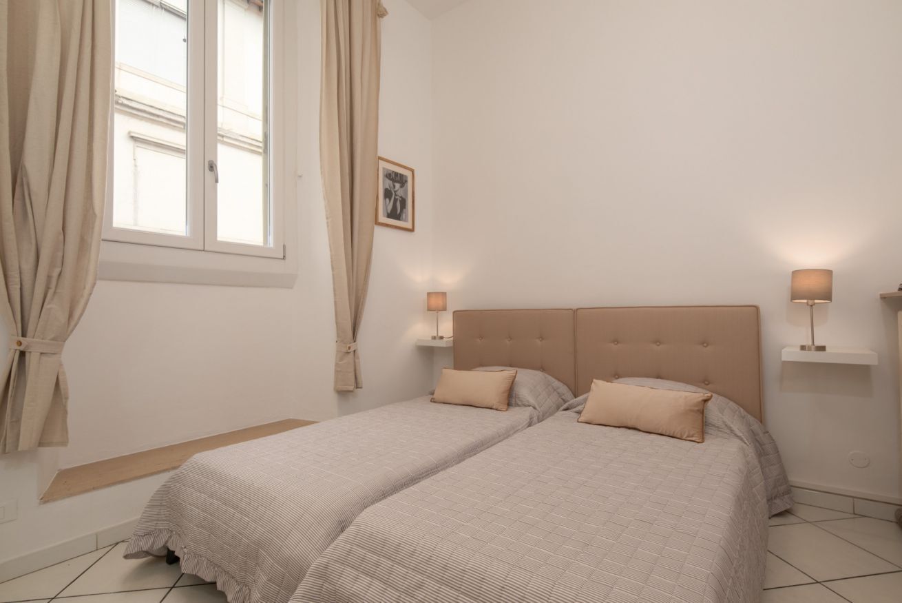 A bedroom with two beds, a window with curtains, bedside lamps, and a framed picture on the wall.