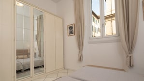 Quarto com cama, guarda-roupa, janela com cortinas e vista para edifícios.