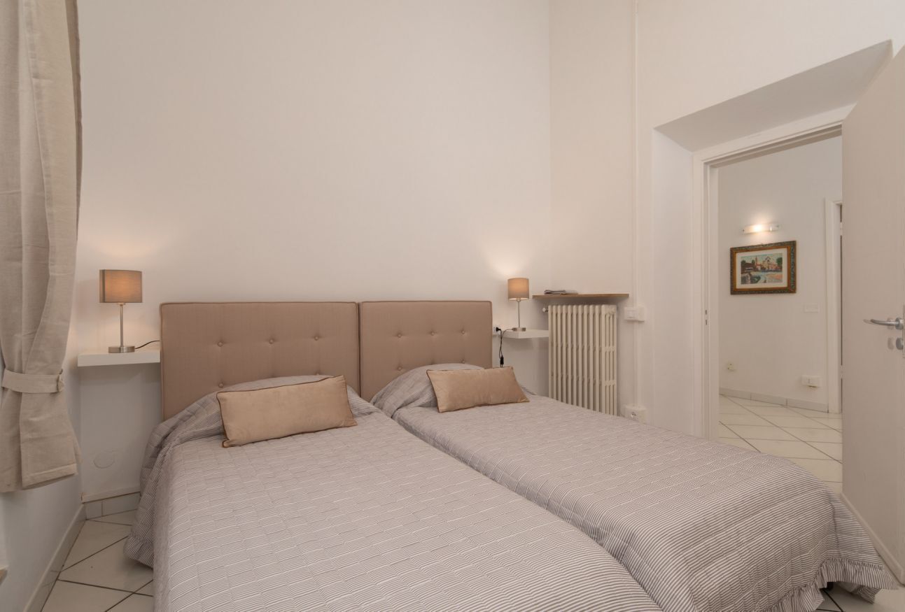 A bedroom with two beds, a headboard, bedside lamps, and a radiator.