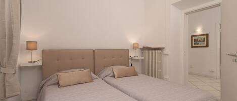 A bedroom with two beds, a headboard, bedside lamps, and a radiator.