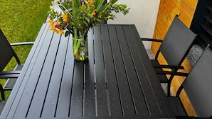 Taula i cadires de metall negre amb coixinets de seients teixits, una planta en test amb flors grogues i una tanca de fusta al fons.