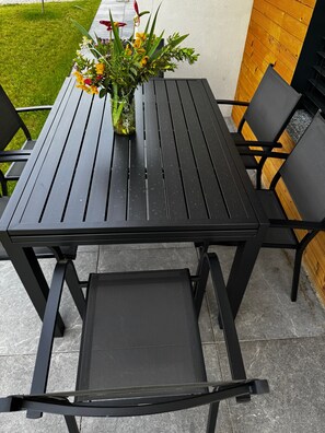 A black metal table and chairs with a woven seat cushion, a potted plant with yellow flowers, and a wooden fence in the background. - Apartment 'Tommaso' in Villa Flaminia, Colico, with Garden and BBQ (Colico)